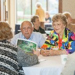A man and woman seated at a table are looking at a woman across the table who is holding a sheet of a paper